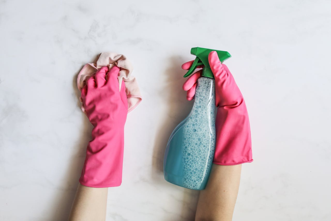 hero-img-01 A pair of hands in pink gloves are cleaning with a spray bottle on a white background.